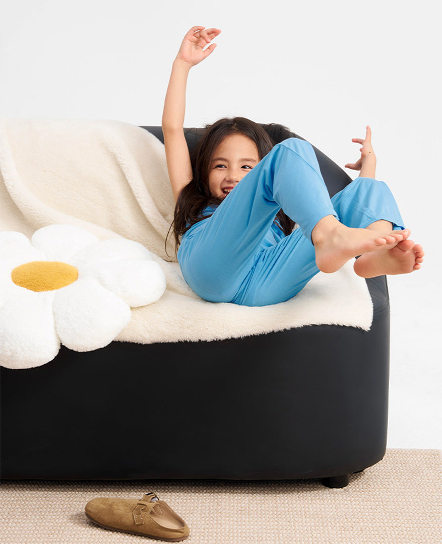 Young girl laughing and playing on a minimalist black sofa, wearing a vibrant azure blue sensory loungewear set from the PYBBLE kids modal loungewear collection. Beside her is a plush white daisy cushion against a clean, bright studio background.